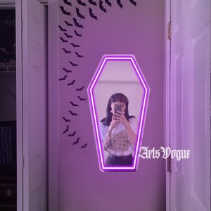 May include: A purple neon coffin-shaped mirror with a person reflected in it. The mirror is hanging on a wall with black bats cutouts. The text "Arts Vogue" is visible in the reflection.