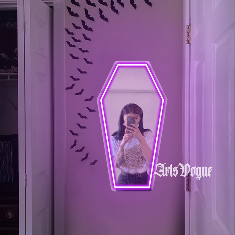 May include: A purple neon coffin-shaped mirror with a person reflected in it. The mirror is hanging on a wall with black bats cutouts. The text "Arts Vogue" is visible in the reflection.