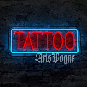 May include: A neon sign with a blue border and red letters that reads "TATTOO". The sign is mounted on a brick wall. The text "Arts Vogue" is written below the sign.