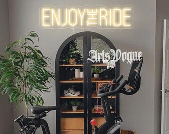 May include: A black and white neon sign that reads "ENJOY THE RIDE" is mounted on a gray wall above a black exercise bike. The bike is in front of a black cabinet with glass doors. The cabinet has shelves with plants and shoes on them. The sign is in a home gym.