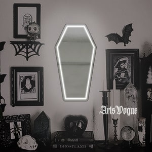 May include: A white coffin-shaped mirror with a white neon outline, hanging on a wall with black and white decor, including a spiderweb, a framed picture of a ghost, and a framed picture of a bat.