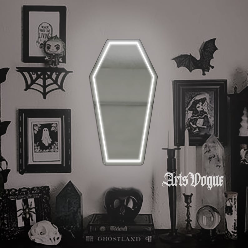 May include: A white coffin-shaped mirror with a white neon outline, hanging on a wall with black and white decor, including a spiderweb, a framed picture of a ghost, and a framed picture of a bat.