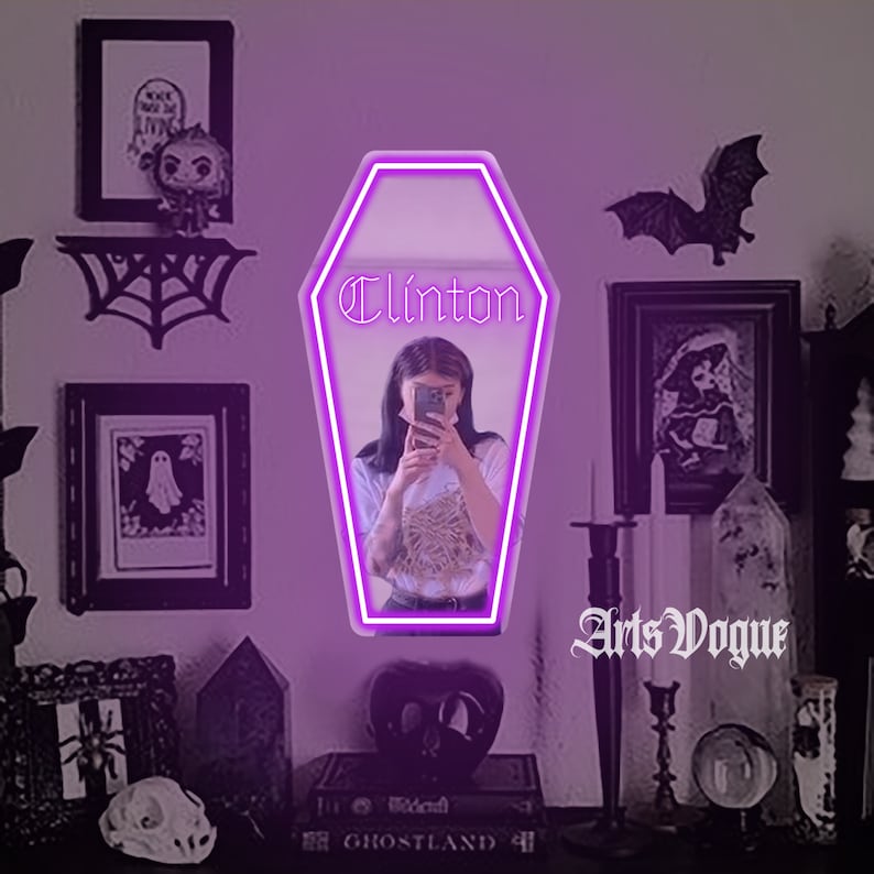 May include: A neon pink coffin-shaped sign with the name "Clinton" in a gothic font. The sign is in front of a woman with dark hair, wearing a white shirt and holding a phone. The background includes a black spiderweb, a framed picture of a ghost, and a book titled "Ghostland".