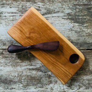 May include: A wooden serving board with a round cutout and a small wooden butter knife. The board is made of light brown wood with a natural grain pattern.