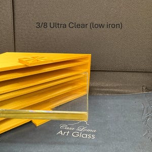 May include: A stack of yellow, folded material with a clear glass sheet in the foreground. The text "3/8 Ultra Clear (low iron)" is visible. The words "Casa Loma Art Glass" are also visible.