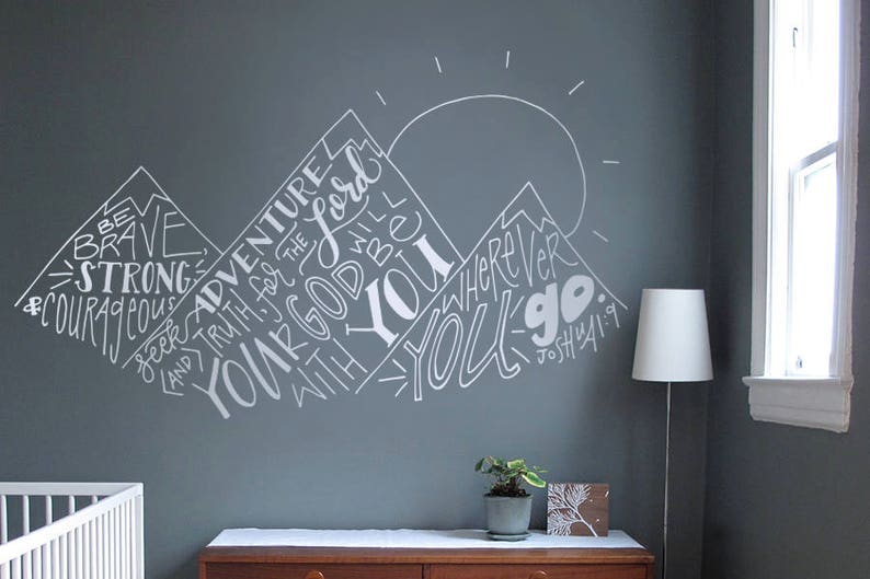 May include: White text on a grey wall reads "Be Brave & Courageous, Seek Adventure, And Truth for the Lord, Your God Will Be With You Wherever You Go." The text is written in a stylised font and arranged in the shape of mountains. The text is accompanied by a small sun graphic.