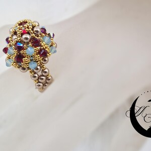 May include: A beaded ring with a cluster of colorful beads. The ring features gold, red, blue, and pearl-colored beads. The ring is on a white surface. A black logo of a peacock is in the bottom right corner.