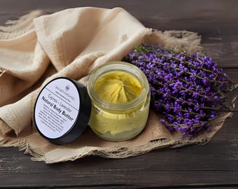 Puede incluir: Un tarro de manteca corporal amarilla, sin tapa, junto a un tarro con tapa negra. Descansan sobre un paño beige, con un manojo de lavanda morada. La etiqueta dice "Cypress - Lavender Natural Body Butter".