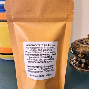 May include: A brown paper pouch with a white label listing the ingredients and instructions for a caffeine-free, vegan herbal tea blend. The ingredients include Tulsi, Fennel, Ashwagandha, Shatavari, Ginger, and Licorice. The instructions state to steep one teaspoon of the blend in boiling water for 7 minutes.