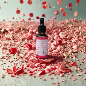 May include: A small amber glass dropper bottle labeled Rose Elixir by Yuliya's Naturals, featuring a black dropper cap. The bottle is surrounded by a dense scattering of pink and red rose petals on a light green surface with petals falling.