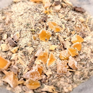 May include: A close-up of a mixture of dried herbs and spices, including pieces of dried ginger. The mixture is a light brown color and has a variety of textures.