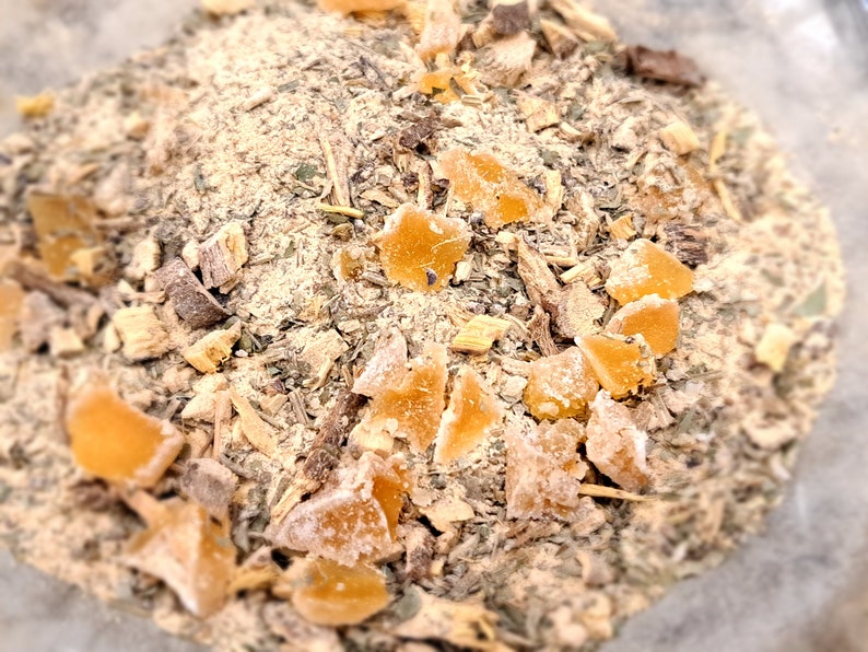 May include: A close-up of a mixture of dried herbs and spices, including pieces of dried ginger. The mixture is a light brown color and has a variety of textures.