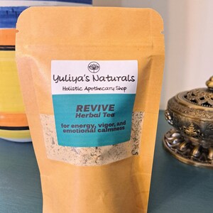 May include: A brown paper pouch with a label that reads "Yuliya's Naturals Holistic Apothecary Shop REVIVE Herbal Tea for energy, vigor, and emotional calmness".