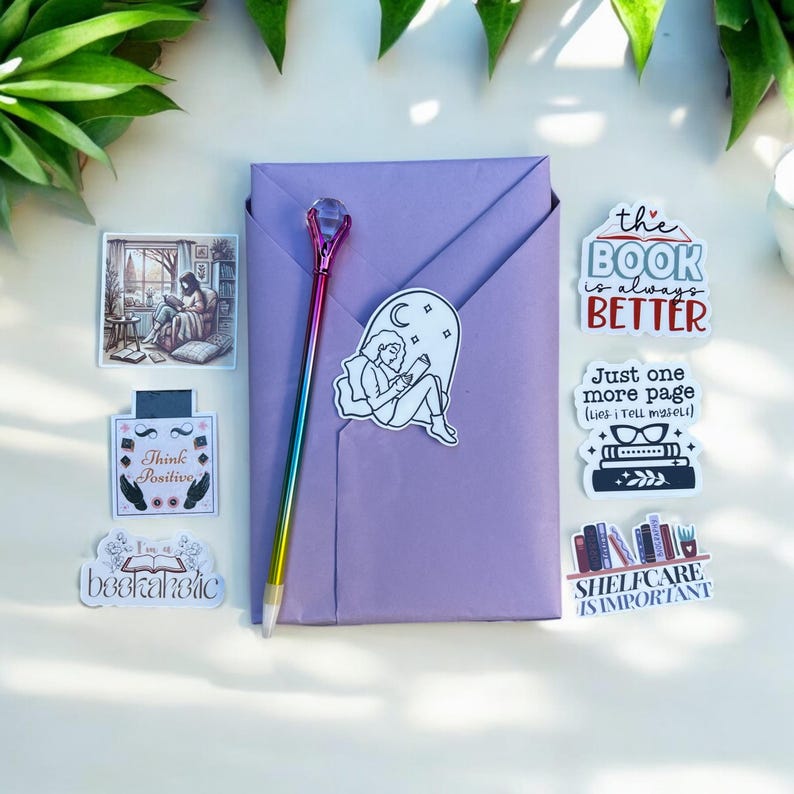 May include: A purple envelope with a rainbow pen and several stickers. The stickers feature illustrations of books, a person reading, and text that says "The book is always better", "Just one more page (lies I tell myself)", "Think Positive", "I am a bookaholic", and "Shelfcare is important".