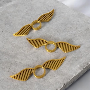 May include: Three golden rings with wings, resembling the Golden Snitch from a popular fantasy series. Each ring has two wings with parallel lines, creating a detailed and textured appearance. The rings are displayed on a light gray surface.