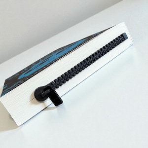May include: A white book with a black zipper bookmark. The book has a blue and black cover design. The zipper bookmark is black and runs along the edge of the book, with the zipper pull at the bottom.