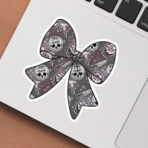 May include: A gray bow-shaped sticker with a repeating pattern of skulls, axes, knives, and the text "STAY FOREST" and "THE HUSBAND DID IT". The sticker has a white border and is placed on a laptop.