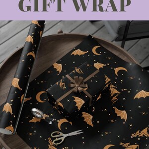 May include: Black gift wrap with a Night Court theme, featuring gold bats, moons, and stars. A wrapped gift with a brown ribbon sits on the wrap, along with a roll of the same paper. Scissors and a silver tape dispenser are also on the table.