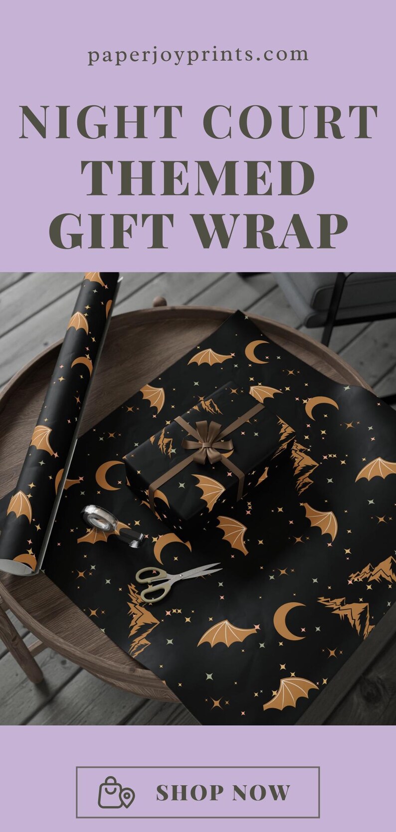 May include: Black gift wrap with a Night Court theme, featuring gold bats, moons, and stars. A wrapped gift with a brown ribbon sits on the wrap, along with a roll of the same paper. Scissors and a silver tape dispenser are also on the table.