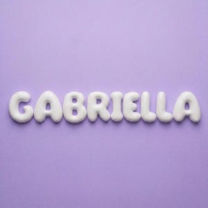 May include: The name GABRIELLA is spelled out in individual, three-dimensional, white, rounded, bubble-style letters. Each letter has a smooth, glossy finish and is arranged in a single horizontal line against a solid, light purple background.