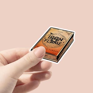 May include: A sticker featuring a book with the title "Iron Flame" in a stylized font. The cover is orange and brown with decorative elements. The author's name, Rebecca Yarros, is also visible.