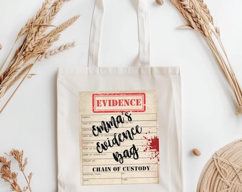 May include: Cream-colored tote bag with a graphic that reads "Emma's Evidence Bag" in black script. The graphic includes a red "Evidence" stamp and a blood spatter design. The bag has two handles.