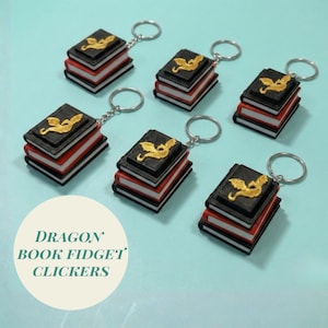 May include: Several dragon book fidget clickers. Each keychain features a miniature book with black covers, red pages, and a gold dragon emblem. The text "DRAGON BOOK FIDGET CLICKERS" is in a white circle.