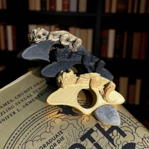 Puede incluir: Tres sujetapáginas con forma de dragón en oro, negro y gris. Cada dragón descansa sobre una base curva con una abertura circular. Los sujetapáginas están encima de un libro con letras doradas.