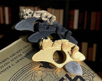 Può includere: Tre fermapagina a forma di drago in oro, nero e grigio. Ogni drago poggia su una base curva con un'apertura circolare. I fermapagina sono sopra un libro con scritte dorate.