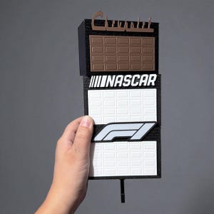 Peut inclure: Un panneau multicouche avec le mot "Chevrolet" en marron, un motif de barre chocolatée, le mot "NASCAR" et le logo F1. Le panneau est tenu par une main sur un fond gris.