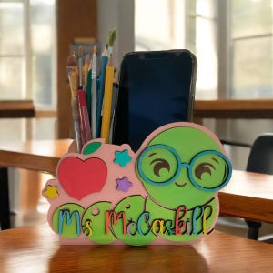 May include: A pink and green wooden desk organizer shaped like a worm with glasses. The organizer has a heart, stars, and a space for a phone. It says "Ms. Mcaskill" on the front.