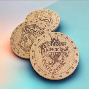 May include: Three round coasters with the crests of Hogwarts houses: Hufflepuff, Slytherin, and Ravenclaw. Each coaster features a house emblem and name in a decorative font, with the letters "HP" around the edge. The coasters are made of a light brown material.