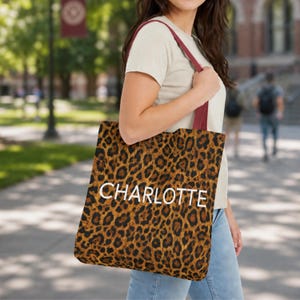 May include: A tote bag featuring a brown and black leopard print pattern with the name CHARLOTTE printed in white capital letters across the center. The bag has two dark red shoulder straps and a square shape for carrying personal items.