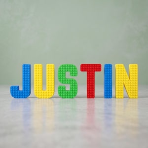 May include: The name JUSTIN spelled out in three-dimensional block letters constructed from small plastic building bricks. The letters J, U, S, T, I, and N are colored blue, yellow, green, red, blue, and yellow respectively, standing upright.
