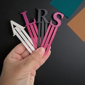May include: A hand holds a collection of letter-shaped bookmarks. The bookmarks are in various colors, including white, pink, and black. The letters are "A", "L", "R", and "S". The background is black with colored paper swatches.