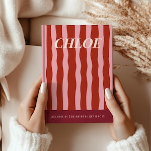 May include: A journal with a red and pink striped cover. The name "CHLOE" is printed in white script across the top. The bottom of the journal reads "ARCHIVE OF SENTIMENTAL ARTIFACTS" in white text. The journal is held by a person.