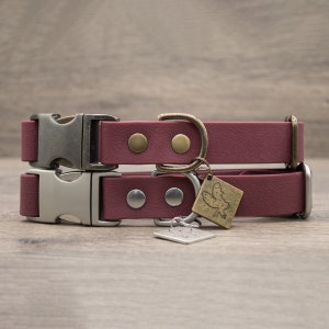 May include: Two burgundy dog collars with metal buckles and hardware. Each collar has a matching metal tag with a decorative design. The collars are stacked on top of each other, showcasing the design and hardware.