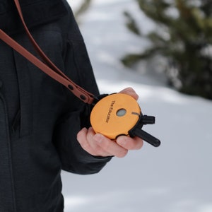 Puede incluir: Un dispositivo electrónico naranja para adiestramiento canino con el texto "The Educator" es sostenido por una persona. El dispositivo tiene una correa de cuero marrón y componentes negros. El fondo es una escena nevada al aire libre.