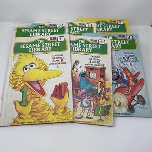 May include: A collection of vintage Sesame Street Library books. The covers feature colorful illustrations of Big Bird, Cookie Monster, and other Muppet characters. The books are labeled with volumes 1, 4, 5, 7, 8, and 9, and feature letters and numbers.