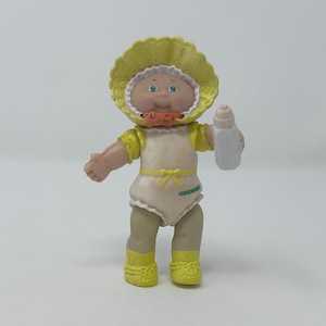 May include: A small plastic doll with a round face, blue eyes, and a yellow bonnet with a white ruffled edge. The doll wears a cream-colored romper with yellow polka dots, a yellow waist tie, and yellow boots, while holding a white baby bottle.