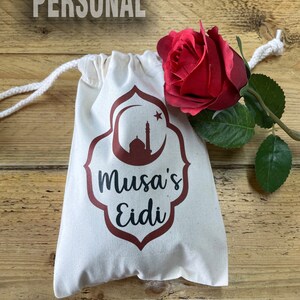 Può includere: Un sacchetto color crema con coulisse, con un disegno bordeaux raffigurante una moschea e la scritta "Musa's Eidi". Una rosa rossa con foglie verdi è appoggiata accanto al sacchetto. Il testo "JUST GOT PERSONAL" è in alto.