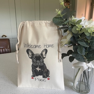 Peut inclure: Un sac en coton crème avec un cordon de serrage. Il présente une illustration d'un bouledogue français noir, avec le texte "Welcome Home" et "Pixie" en dessous, encadré de deux cœurs rouges. Le sac a un aspect naturel et rustique.