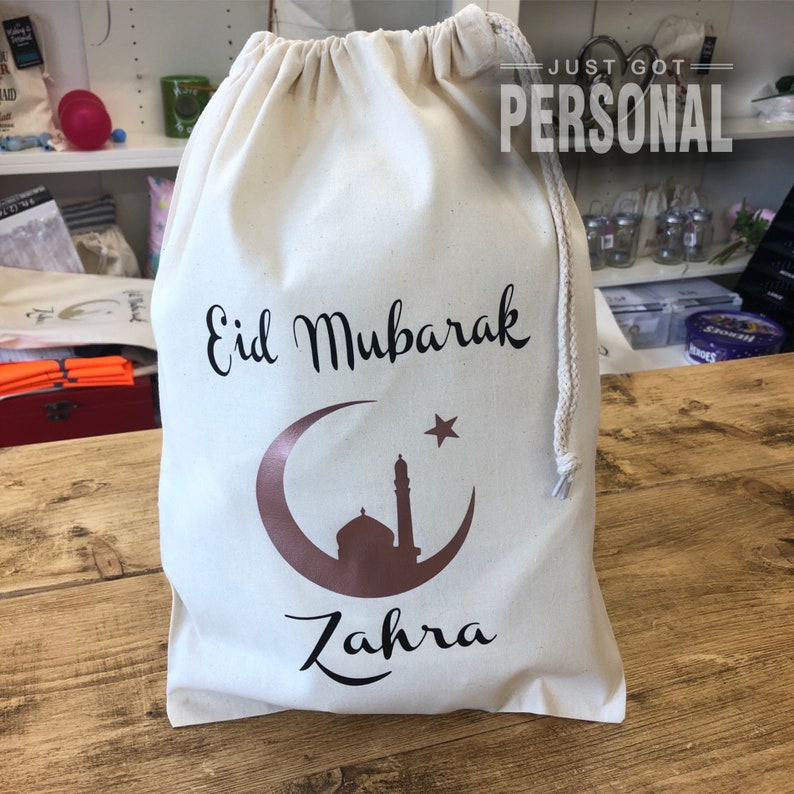May include: A white drawstring bag with the text "Eid Mubarak" and a gold crescent moon and star design. The bag also has the name "Zahra" printed on it.