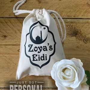 Op de afbeelding: Een beige trekkoordtas met een zwart ontwerp van een maan, ster en moskee, met de tekst "Zoya's Eidi". De woorden "JUST GOT PERSONAL" staan eronder. Naast de tas ligt een witte roos.