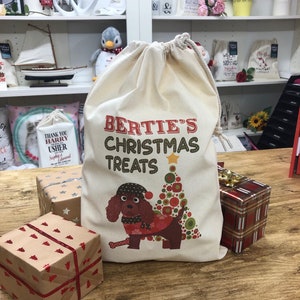 May include: A cream-coloured drawstring bag printed with "BERTIE'S CHRISTMAS TREATS" and a cartoon dog in festive attire next to a Christmas tree. The bag sits on a wooden table with wrapped presents.