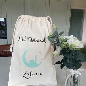 May include: A white drawstring bag with the text "Eid Mubarak" and a crescent moon with a star and a mosque inside. The bag also has the name "Zaheer" printed on it.