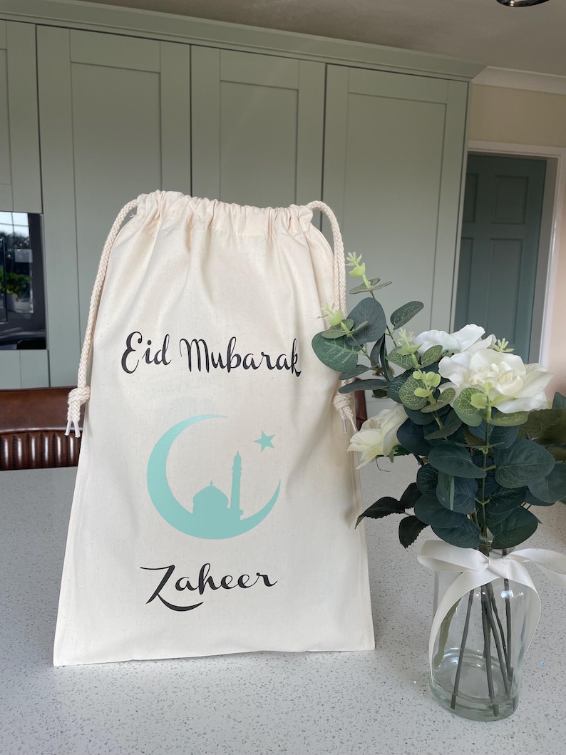 May include: A white drawstring bag with the text "Eid Mubarak" and a crescent moon with a star and a mosque inside. The bag also has the name "Zaheer" printed on it.