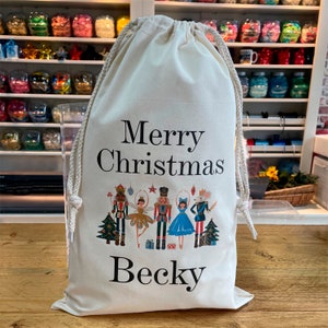 Könnte beinhalten: Ein cremefarbener Geschenkbeutel mit Kordelzug mit dem Text "Merry Christmas" und dem Namen "Becky" in Schwarz. Die Tasche zeigt eine farbenfrohe Illustration von Nussknacker-Figuren und Weihnachtsbäumen.