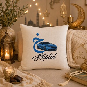 May include: A square beige throw pillow featuring a blue Arabic letter, a blue sports car graphic, and the name Khalid written in black cursive script. The pillow has a textured fabric surface and is centered in the frame.
