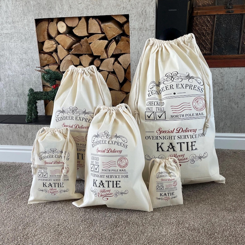 May include: A set of five off-white drawstring canvas bags in varying sizes. Each bag features black and red printed text reading Reindeer Express, Special Delivery, Checked Once Twice, North Pole Mail, and the name Katie.
